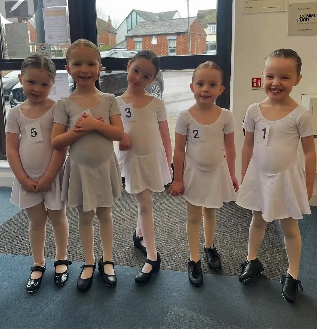 Five young tap dancers in white leotards and tap shoes, smiling in a line
